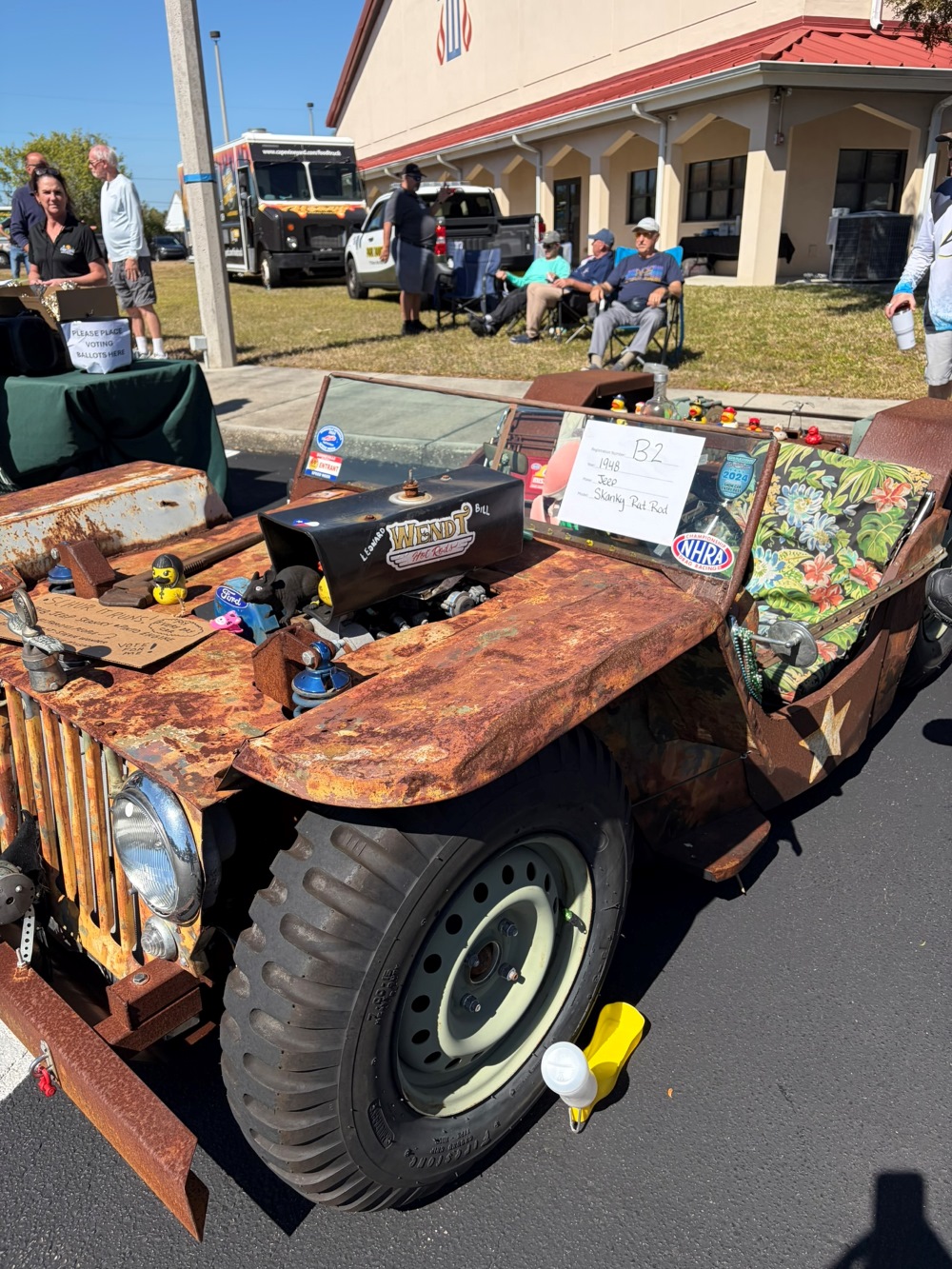 Rusty rat-rod style jeep with exposed engine and novelty decorations parked in front of spectators.